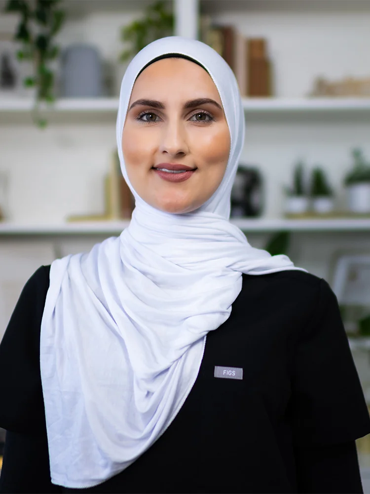 Headshot of Asseya wearing a black scrub top and a hijab, standing against a cabinet with some plants blurred background. Their attire suggests a professional setting, possibly in the medical or dental field.