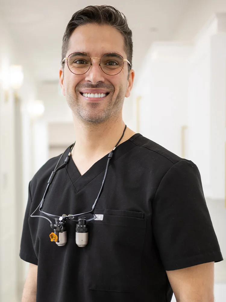Headshot of Dr. Bakkali wearing a black scrub top and dental loupes around their neck, standing against a blurred hallway background.