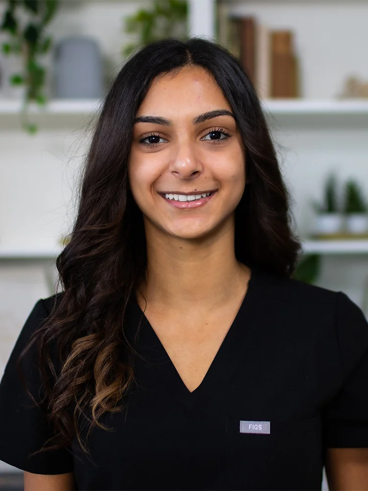 Headshot of Kaila wearing a black scrub top and standing against a cabinet with some plants blurred background. Their attire suggests a professional setting, possibly in the medical or dental field.