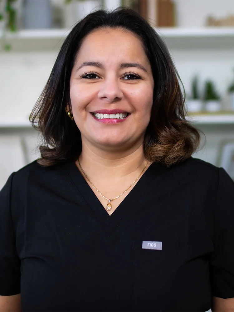 Headshot of Sandra wearing a black scrub top and standing against a cabinet with some plants blurred background. Their attire suggests a professional setting, possibly in the medical or dental field.