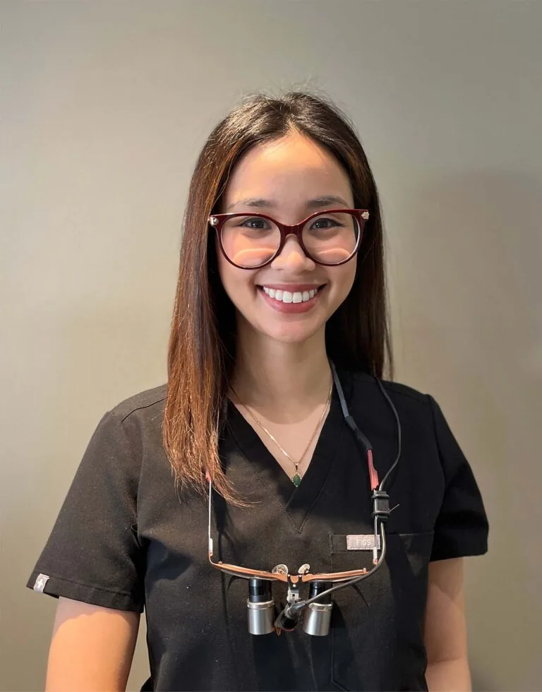 Headshot of Dr. Ha wearing a black scrub top and dental loupes around their neck, standing against a plain background.